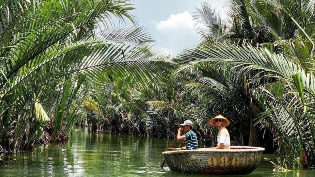 Two people ride a round basket boat through lush green palm trees on a calm Hoi An river under a bright, partly cloudy sky.