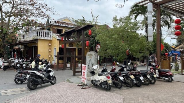 Scooters parked in front of yellow Hoi An buildings with red lanterns hanging in a busy outdoor area.