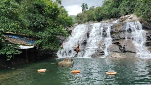 A person stands on rocks under a waterfall near Hoi An, surrounded by greenery and floating orange life rings in the water.