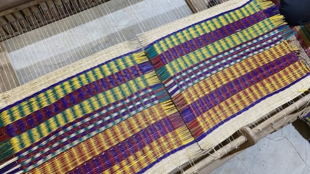 Colorful woven textile in progress on a wooden loom in Hoi An, with hands working at the fabric’s edge.