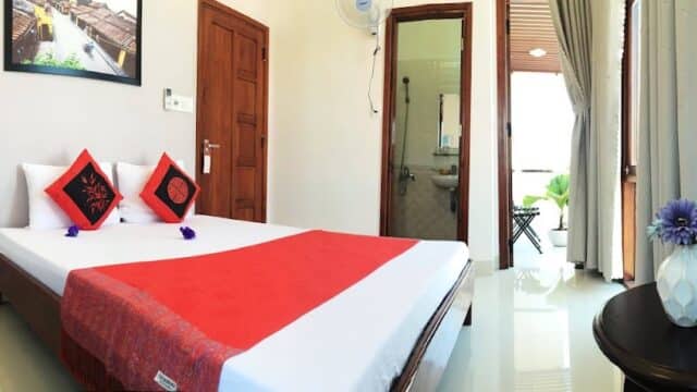 Bright Hoi An bedroom with a double bed, red accents, flowers, and an open door to a balcony and bathroom.