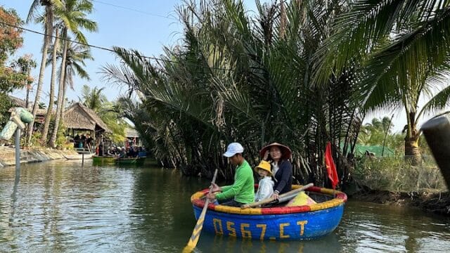 Two people paddle a round blue Hoi An boat with yellow letters through a calm, palm-lined river under a clear blue sky.