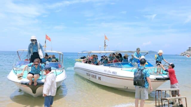 People disembark from two speedboats onto a sandy Hoi An beach, enjoying the sunny day, clear blue sky, and calm sea.