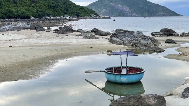 A round blue boat with an umbrella floats in a shallow tidal pool on a rocky Hoi An beach with green hills in the background.
