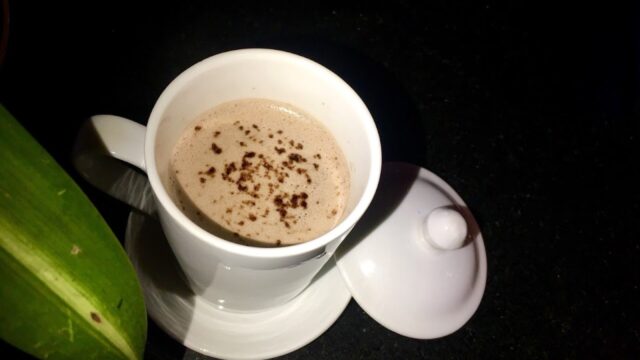 A white cup of coffee with chocolate sprinkles sits on a saucer, evoking cozy Hoi An vibes on a dark surface.