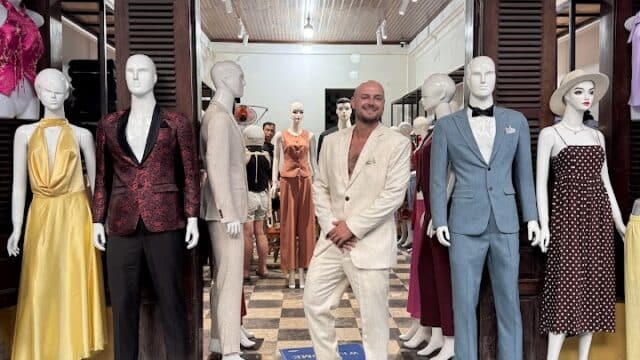 A man in a light suit stands among mannequins outside a Hoi An tailor shop showcasing elegant formal dresses and suits.