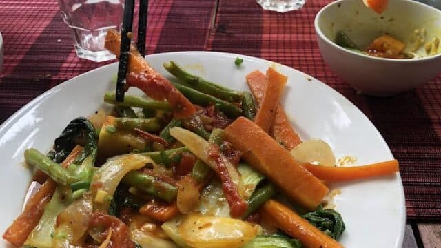 A hand with chopsticks picking Hoi An-style stir-fried vegetables from a white plate on a red table mat.