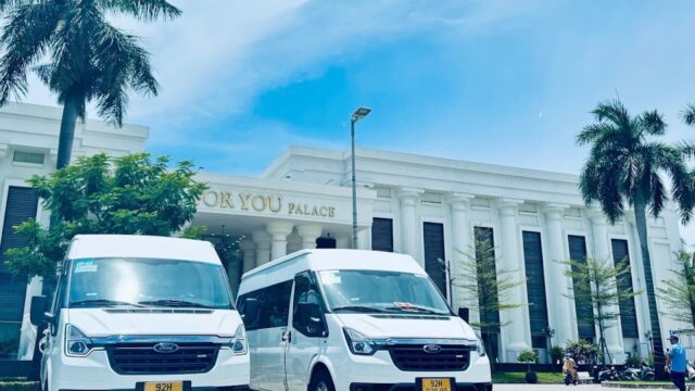 Two white vans parked in front of a white building with columns and palm trees in sunny Hoi An.