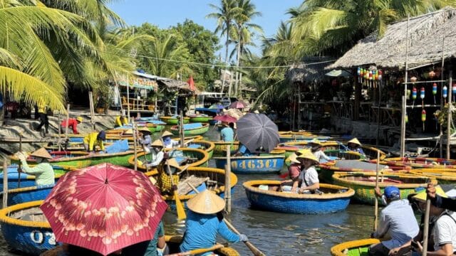 People in round boats with colorful umbrellas paddle on a Hoi An river, surrounded by palm trees and wooden huts under a sunny sky.