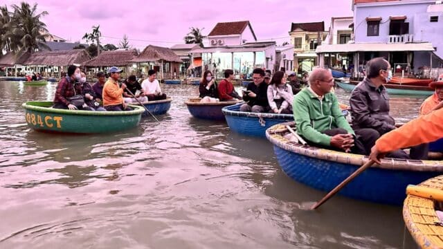 People sit in round basket boats on a river at dusk in Hoi An, with buildings and palm trees beneath a purple sky.