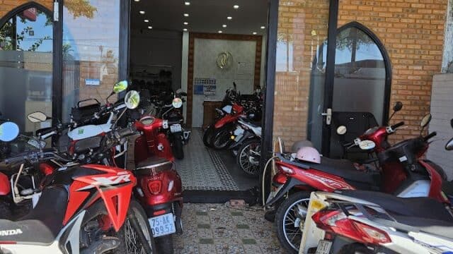 Several motorbikes are parked outside a brick building with glass doors at the entrance in sunny Hoi An.