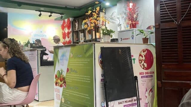 Indoor cafe with a colorful counter, Hoi An-style decorations, menu signs, and customers at tables. Bright and lively atmosphere.