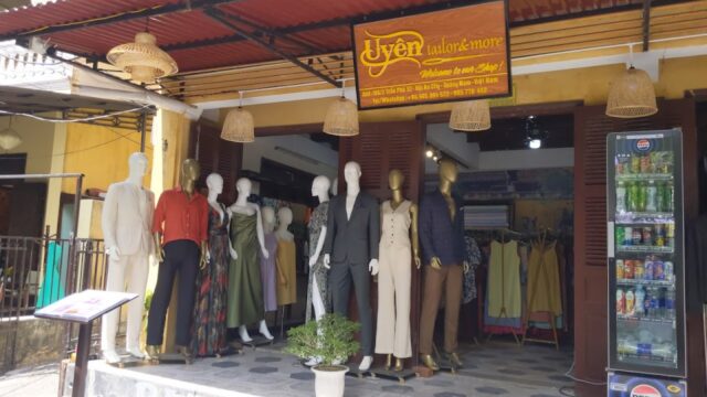 Mannequins in formal and casual wear outside a Hoi An tailor shop, with a drinks fridge to the right.