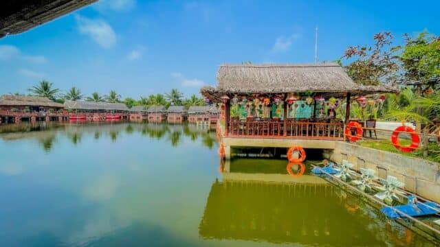 Thatched-roof pavilion over a pond with life rings in Hoi An, surrounded by palm trees under a clear blue sky.