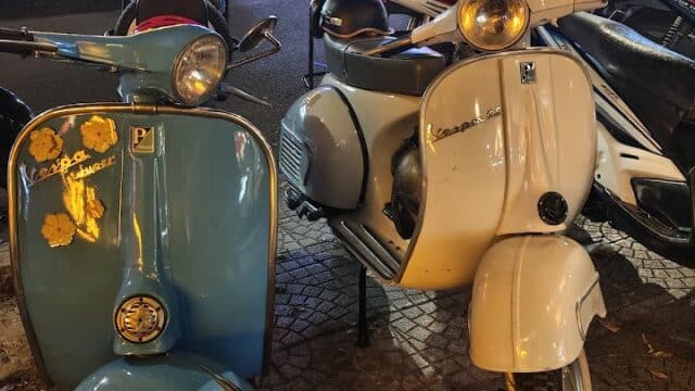 Two vintage Vespa scooters in Hoi An, one blue with yellow stickers and one white, parked on a city street at night.