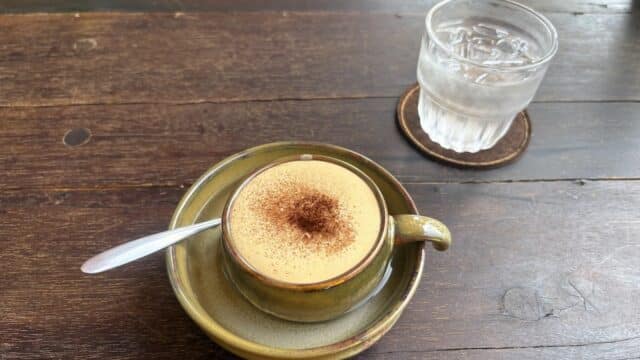 A cup of coffee with foam and cinnamon beside a glass of water sits on a wooden table in charming Hoi An.