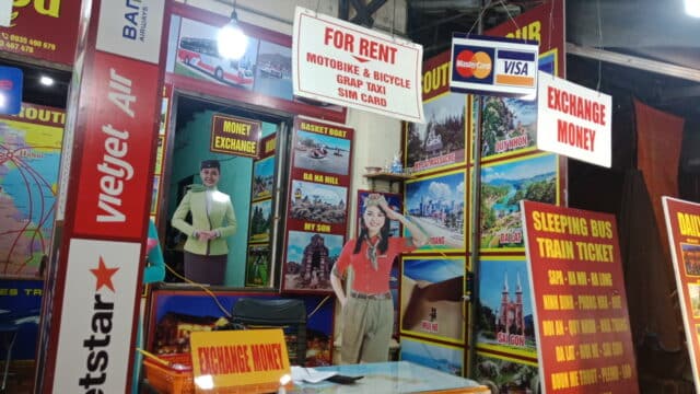 Travel agency booth in Hoi An with signs for rentals, money exchange, SIM cards, and ticket services in a colorful setting.