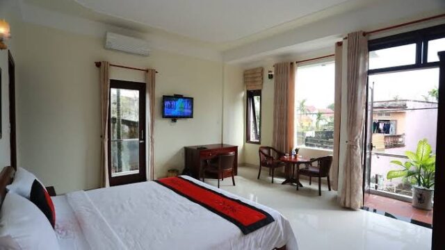 Bright Hoi An hotel room with a bed, desk, chairs, large windows, and a view of a balcony with plants outside.