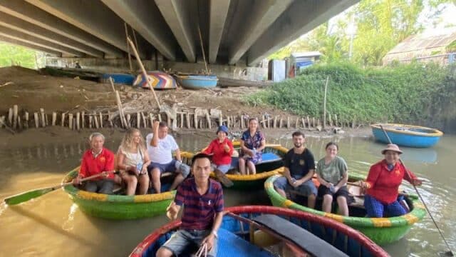 A group of people sit in round boats under a bridge on a river in Hoi An, surrounded by trees and wooden fences.