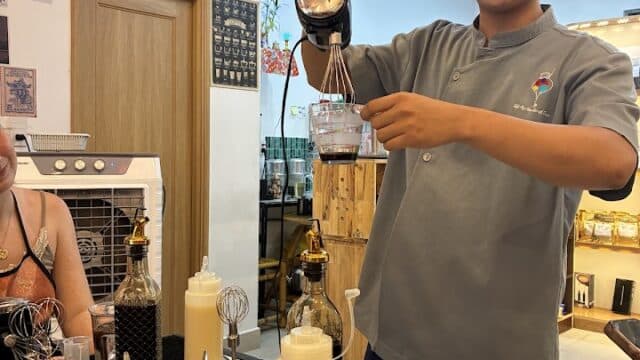 A smiling person in a gray shirt prepares a drink with a mixer in a kitchen, evoking the vibrant spirit of Hoi An.