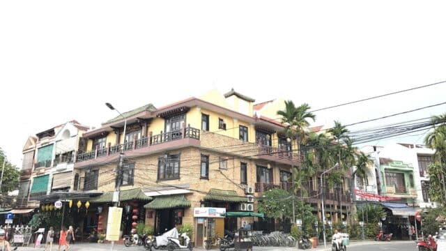 A street corner in Hoi An with a yellow building, shops, parked motorbikes, palm trees, and people walking in an urban area.