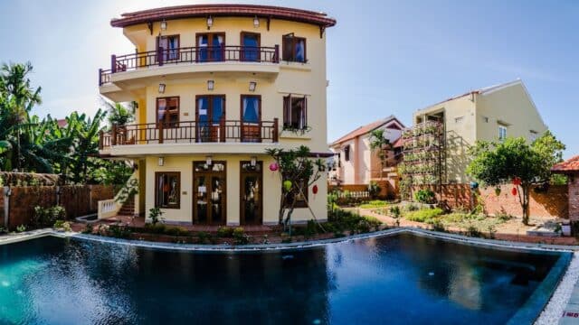 A three-story yellow house with balconies overlooks a large swimming pool in Hoi An, surrounded by plants and neighboring homes.