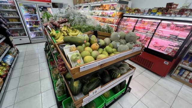 Fresh fruits and vegetables displayed in a grocery store aisle in Hoi An, with coolers and shelves in the background.