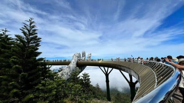 Curved pedestrian bridge held by giant stone hands near Hoi An, with people walking and trees below a blue sky.