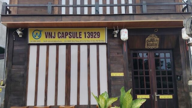 A wooden two-story building in Hoi An with a VNJ CAPSULE 13929 sign, potted plants, stools, and hanging lanterns outside.