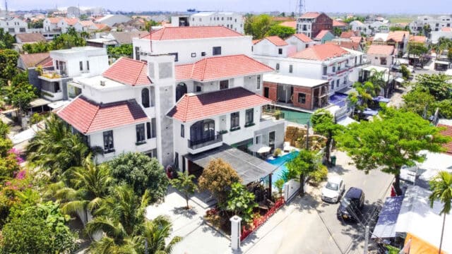 Aerial view of a white villa with red-tiled roofs in a Hoi An suburban neighborhood dotted with trees and parked cars.