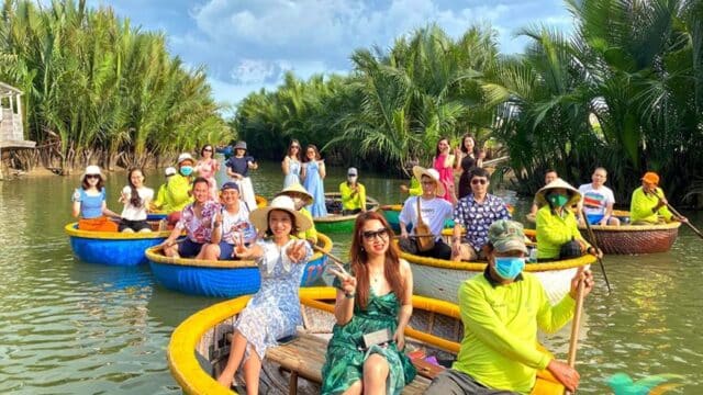People riding colorful round boats on a river in Hoi An, surrounded by lush green palm trees under a partly cloudy sky.