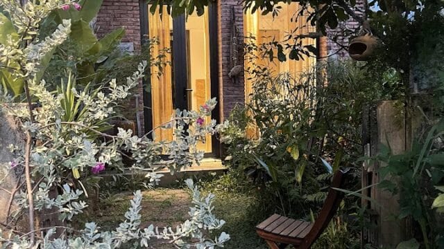 A small Hoi An garden with lush plants, a wooden chair, and a glowing doorway in the background at dusk.