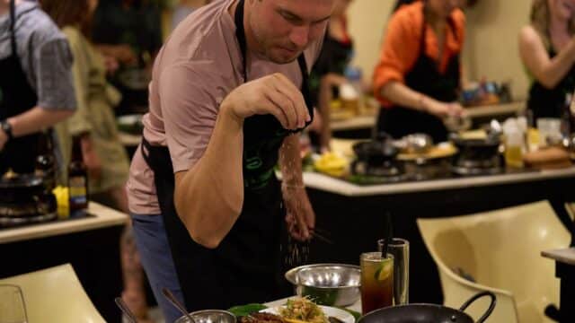 A man sprinkles seasoning on a dish in a busy Hoi An cooking class, with others preparing food in the background.