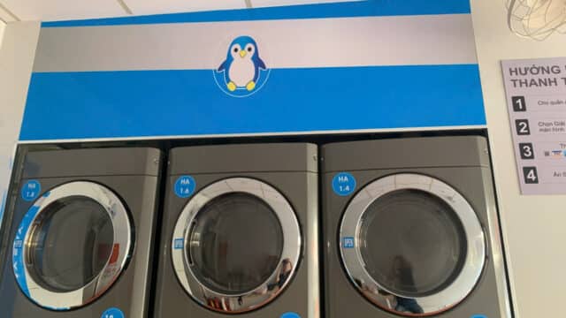 Three front-loading washing machines in Hoi An sit beneath a blue sign with a cartoon penguin and Vietnamese instructions on the right wall.