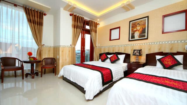 Bright Hoi An hotel room with two double beds, red accents, a table with chairs, large windows, and a painting on the wall.