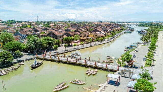 A riverside town with brown-tiled roofs, a bridge, boats on the water, and greenery beneath Hoi An’s clear blue sky.