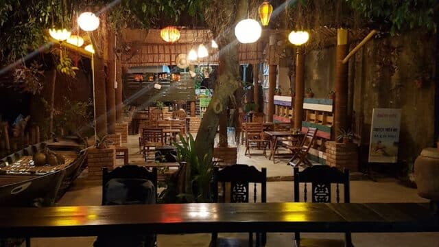 Outdoor restaurant at night in Hoi An with hanging lanterns, wooden tables, and chairs under trees, creating a cozy, inviting atmosphere.