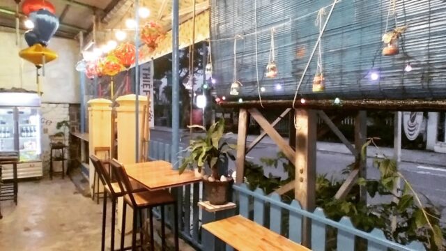 A cozy cafe patio in Hoi An with wooden tables, high chairs, string lights, and potted plants beside a street at night.