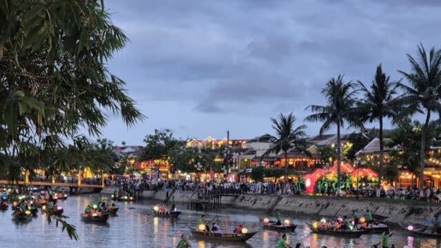 Boats with lanterns float on Hoi An's river at dusk, with crowds and colorful lights along the shore.