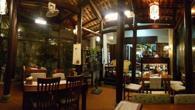 Cozy Hoi An restaurant with wooden tables, warm lighting, glass walls, and lush plants creates an inviting, intimate atmosphere.
