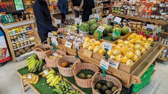 People shop for fruit in a grocery store, reminiscent of Hoi An markets, with baskets of bananas, avocados, melons, and fresh produce on display.