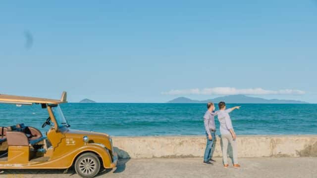 Two people stand by a vintage car near the sea in Hoi An, one pointing towards the horizon under a clear blue sky.