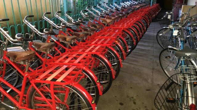 Rows of red bicycles with brown seats are closely lined up in a covered outdoor area beside a metal fence in Hoi An.