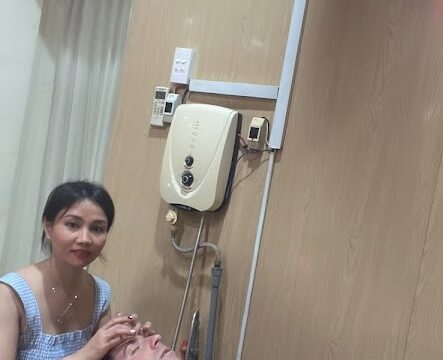 A woman in Hoi An performs a facial treatment on a man lying on a bed in a clinic room with beige walls.