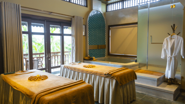 Luxury spa room in Hoi An with two massage tables, robes, a soaking tub, glass shower, and large windows with flowing curtains.