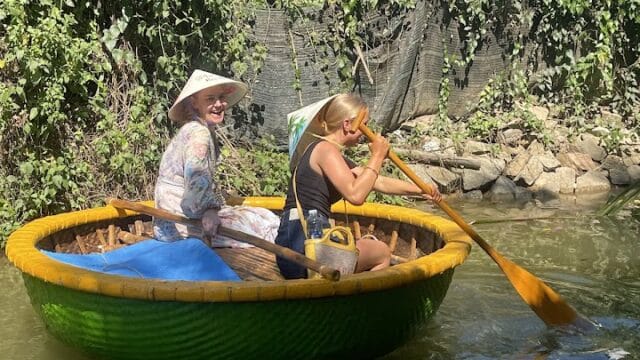 Two women paddle a round, green basket boat on a calm river in Hoi An, surrounded by lush greenery and a stone-lined bank.